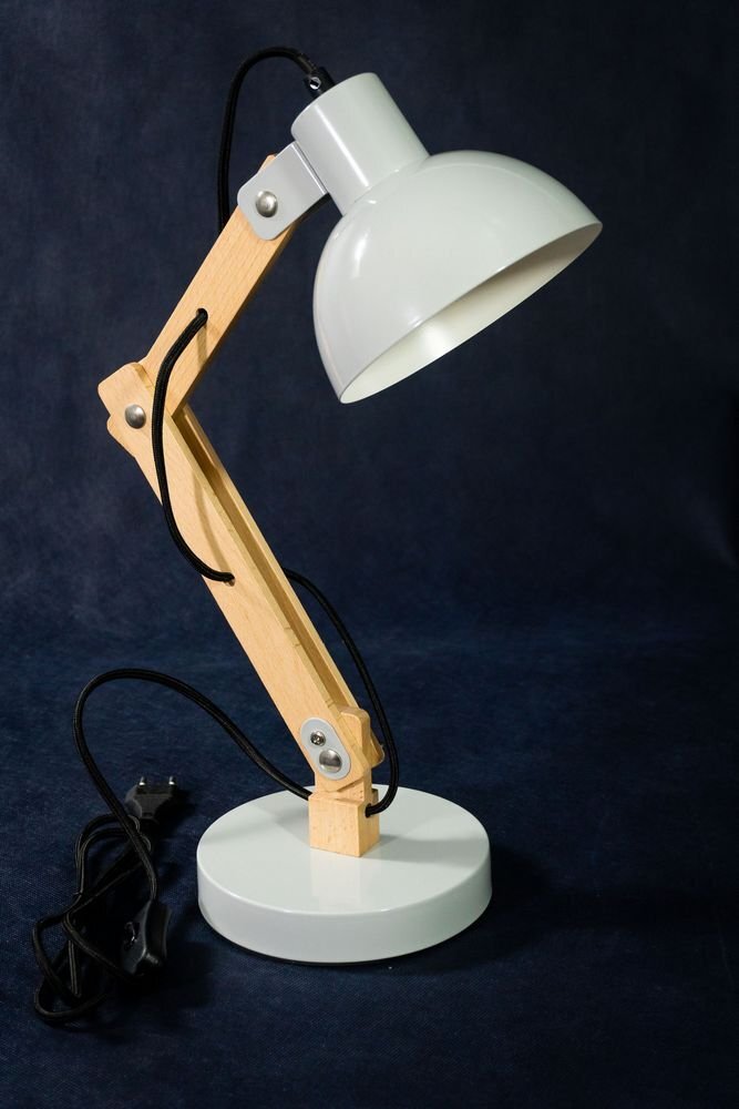 Na ciemnym tle znajduje się biała lampa biurkowa z drewnianym wysięgnikiem i okrągłą podstawą. Kabel z wtyczką jest widoczny obok lampy.
