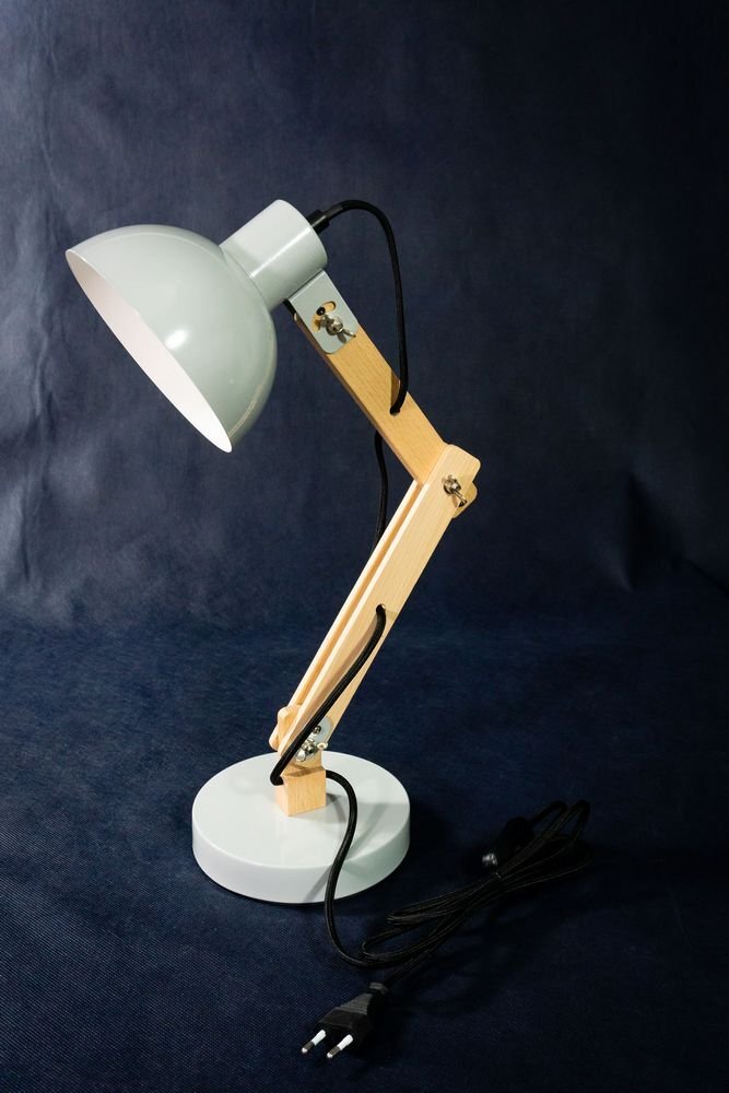 Biała lampa biurkowa na wysięgniku z drewnianymi elementami, stojąca na okrągłej podstawie. Posiada czarny kabel z wtyczką i wyłącznikiem.