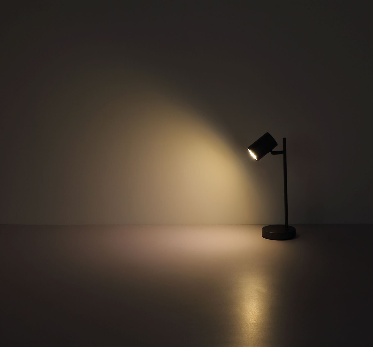 Na obrazie widoczna jest czarna lampa biurkowa emitująca ciepłe światło na ścianę i powierzchnię stołu. Lampa ma prosty, nowoczesny design z cylindrycznym kloszem.