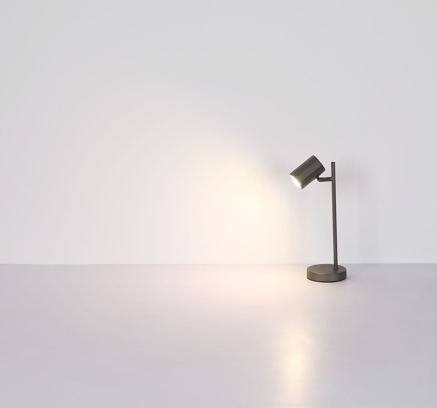Na obrazie widać nowoczesną lampę biurkową o prostym designie, emitującą światło na jasnym tle. Lampa ma cylindryczny klosz i stoi na okrągłej podstawie.