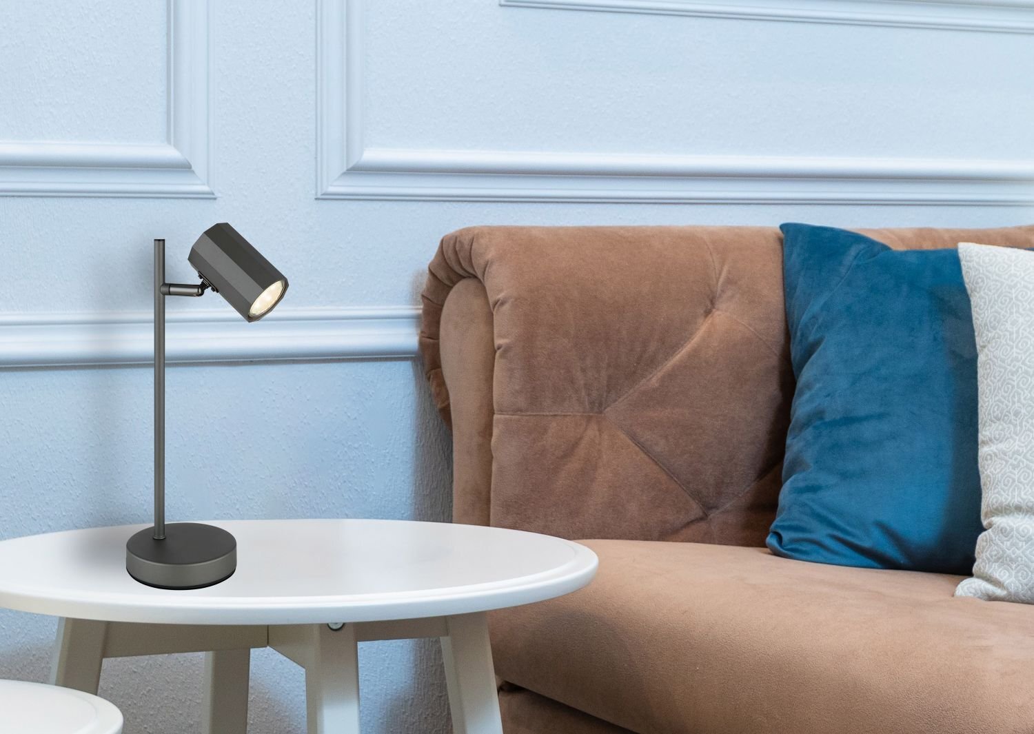 Na białym stoliku znajduje się czarna lampa biurkowa z cylindrycznym kloszem. W tle widoczna jest brązowa sofa z niebieskimi i jasnymi poduszkami.