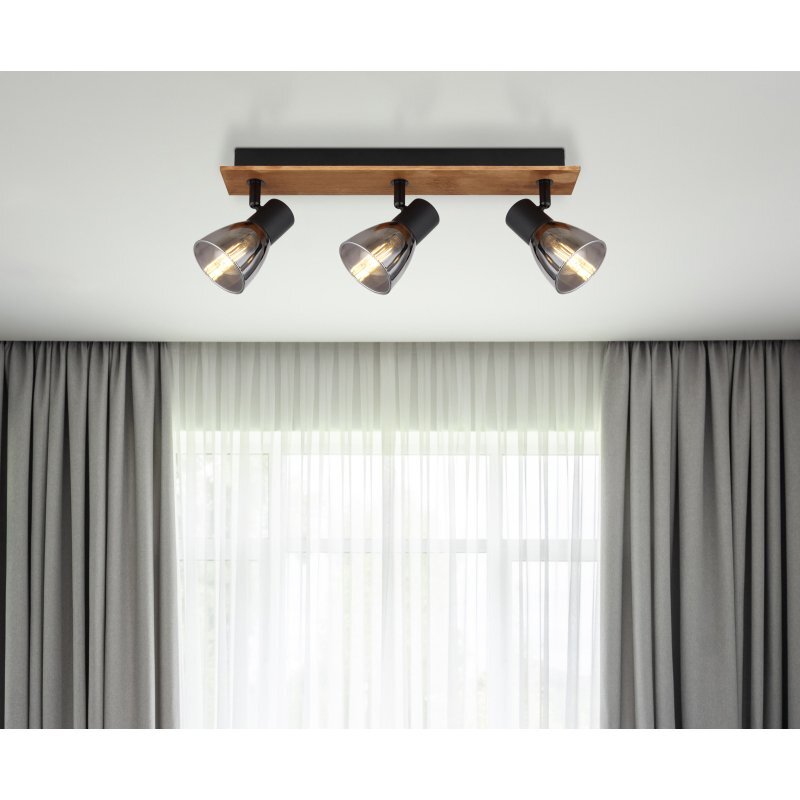 Na suficie zamontowana jest lampa sufitowa z trzema reflektorami, wykonana z drewna w kolorze brązowym i czarnego matu, z przydymionymi szklanymi kloszami. W tle widoczne są zasłony i okno z białą firanką.