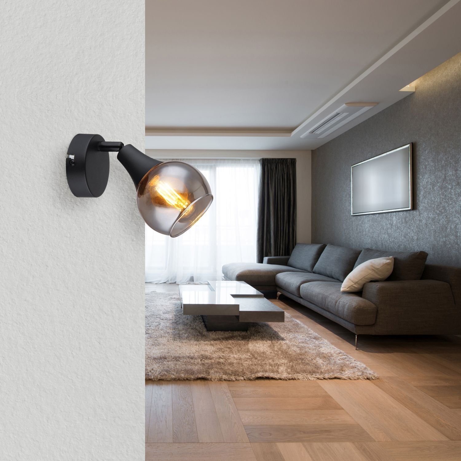 Na ścianie zamontowana jest nowoczesna lampa z czarnym uchwytem i przezroczystym kloszem, w którym widać żarówkę. W tle znajduje się elegancki salon z dużą sofą, stolikiem i dywanem na drewnianej podłodze.