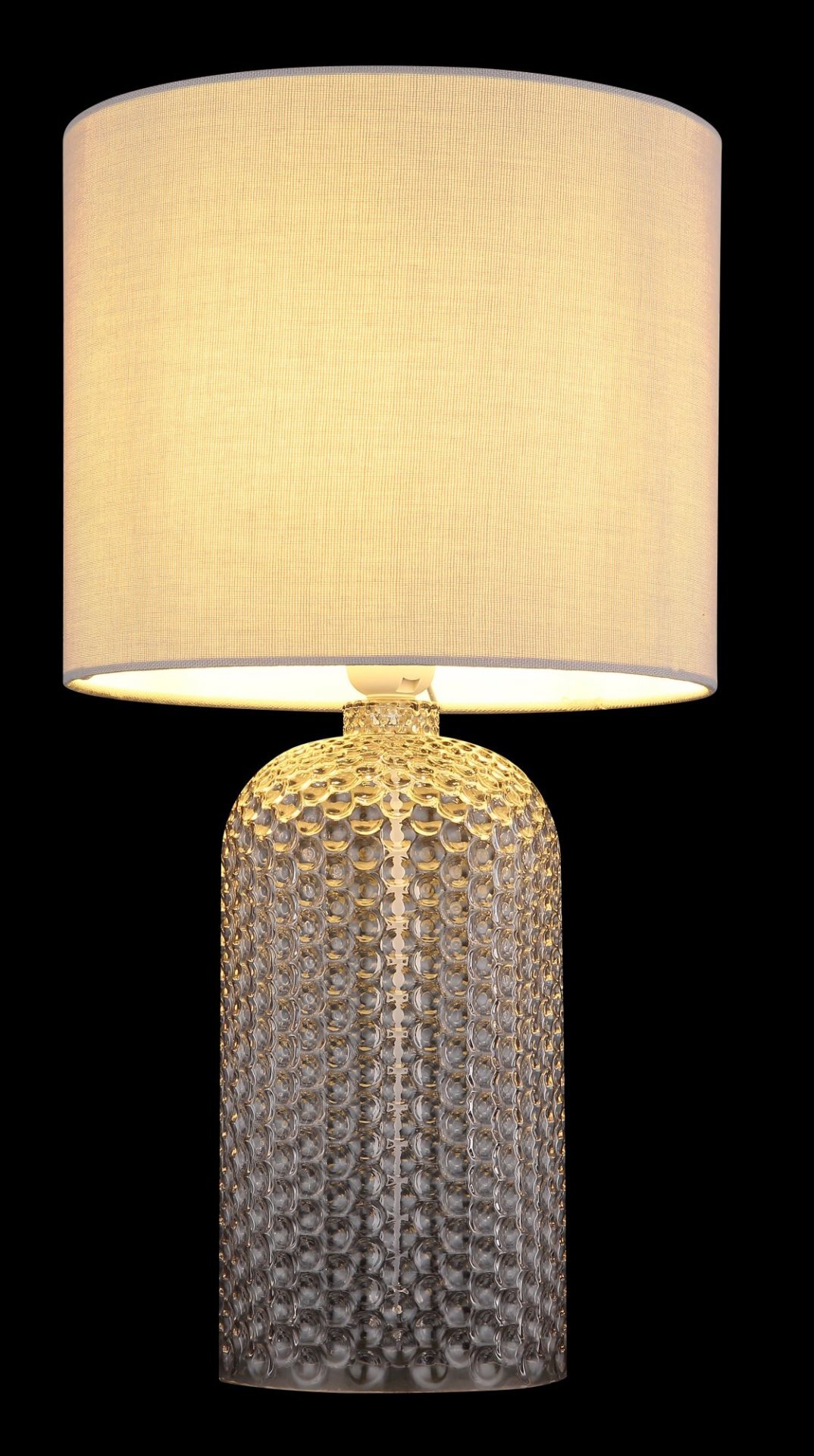 Lampa stołowa z jasnym, cylindrycznym abażurem i ozdobną, przezroczystą podstawą o fakturze przypominającej bąbelki. Światło lampy jest ciepłe i rozproszone.