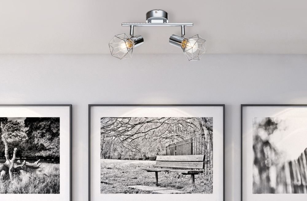 Na suficie zamontowana jest chromowana lampa sufitowa z dwoma reflektorami o geometrycznych kloszach. W tle widoczne są trzy czarno-białe fotografie przedstawiające krajobrazy z ławką i drzewami.