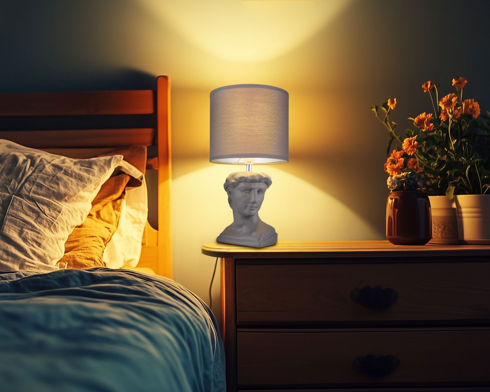Na drewnianej szafce nocnej stoi lampa stołowa z abażurem i podstawą w kształcie popiersia. Obok lampy znajdują się doniczki z kwiatami, a w tle widać łóżko z pościelą.