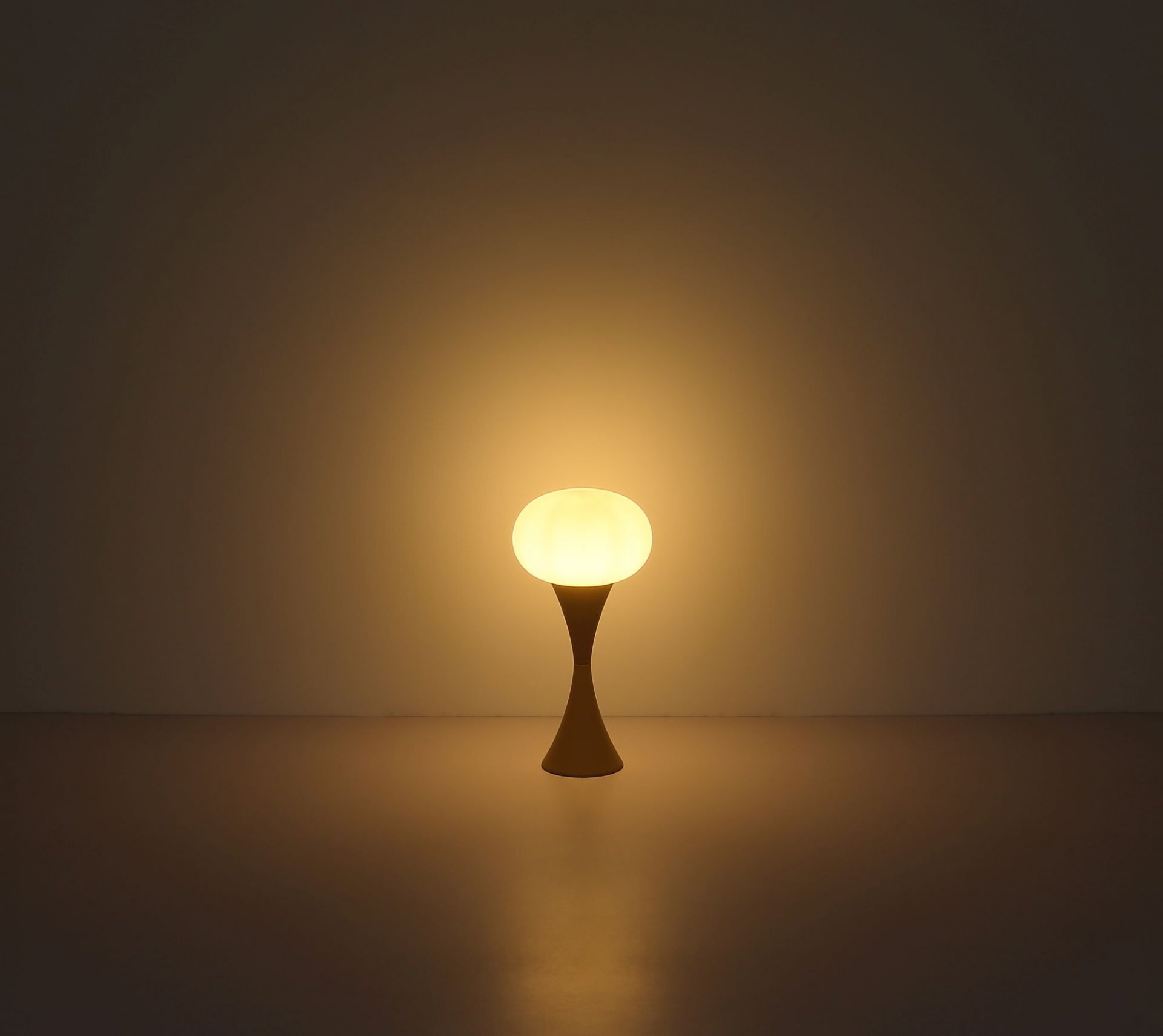 Na obrazie widoczna jest lampa stołowa o nowoczesnym designie, emitująca ciepłe, żółte światło. Lampa ma zaokrąglony klosz i smukłą podstawę, stojącą na gładkiej powierzchni.