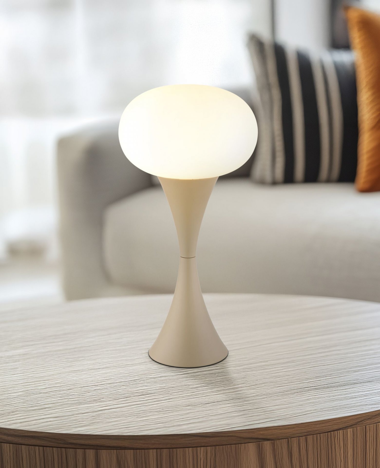 Na okrągłym stoliku znajduje się lampa stołowa o nowoczesnym designie z białym, kulistym kloszem i smukłą podstawą. W tle widoczna jest część kanapy z poduszkami w paski.