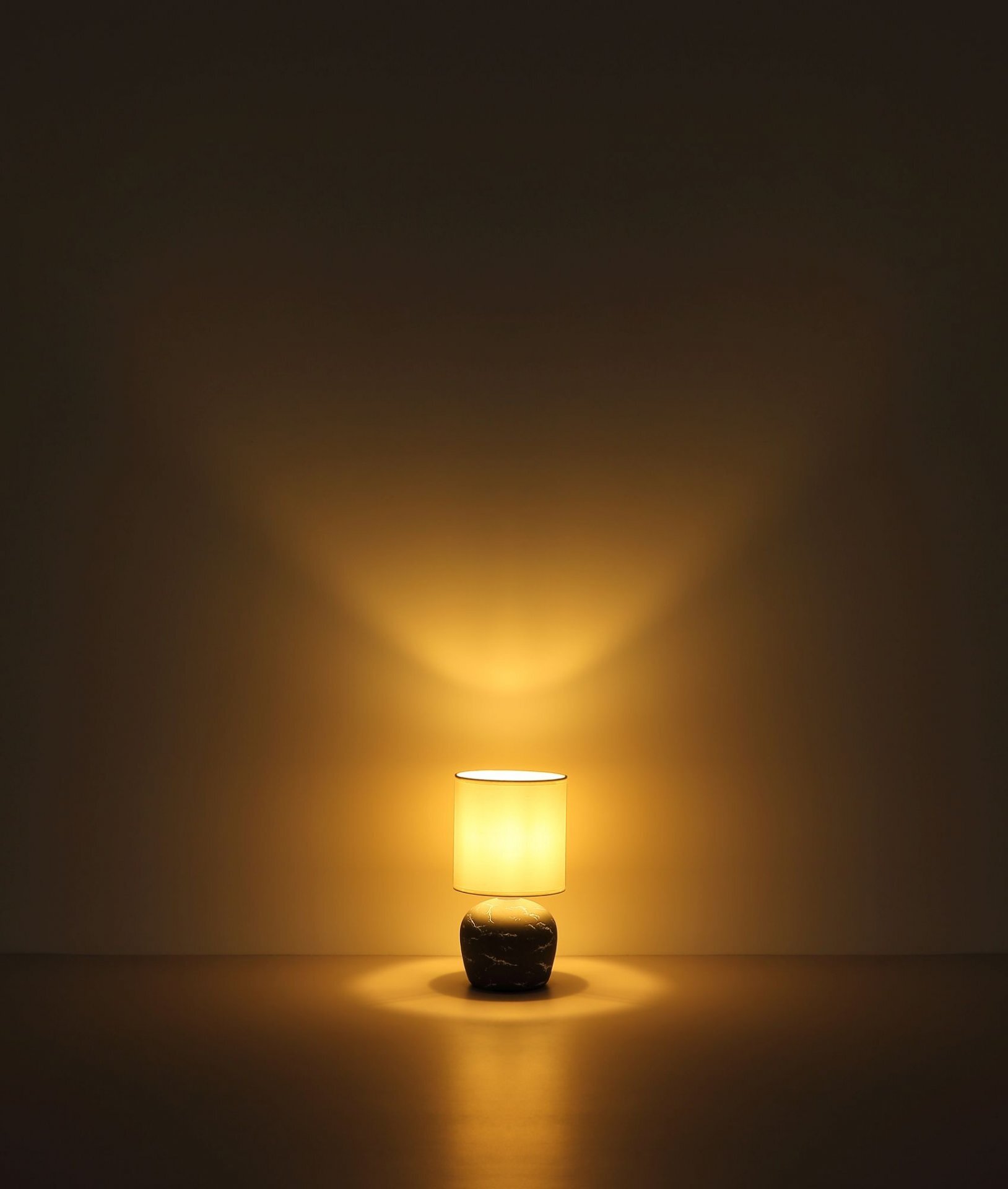 Na obrazie widoczna jest włączona lampa stołowa z jasnym kloszem, emitująca ciepłe, żółte światło. Podstawa lampy ma ciemny, marmurowy wzór.