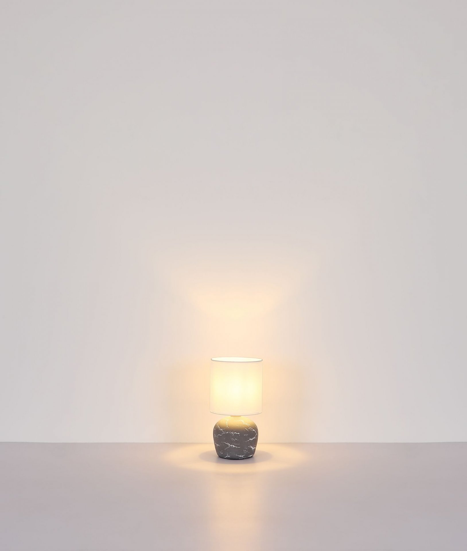 Na obrazie widoczna jest lampa stołowa z jasnym kloszem i podstawą o marmurowym wzorze. Lampa emituje ciepłe światło, które rozprasza się na ścianie i powierzchni, na której stoi.