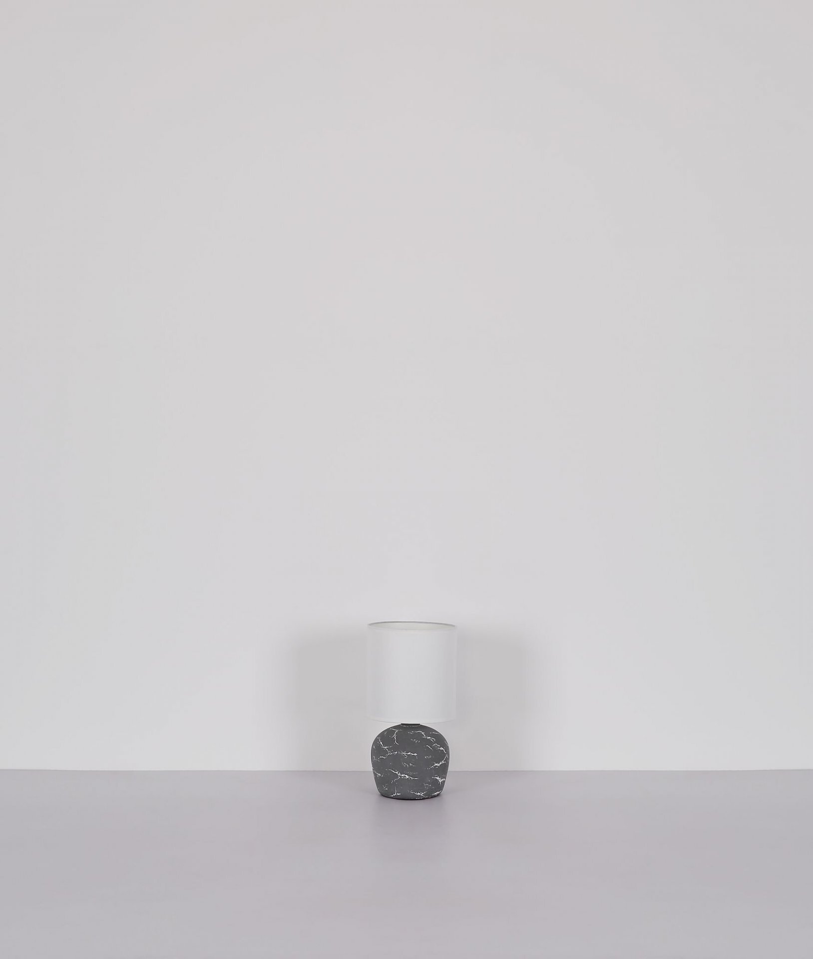 Na obrazie widoczna jest lampa stołowa z białym kloszem i podstawą o marmurowym wzorze, ustawiona na gładkiej powierzchni przed jasnym tłem.