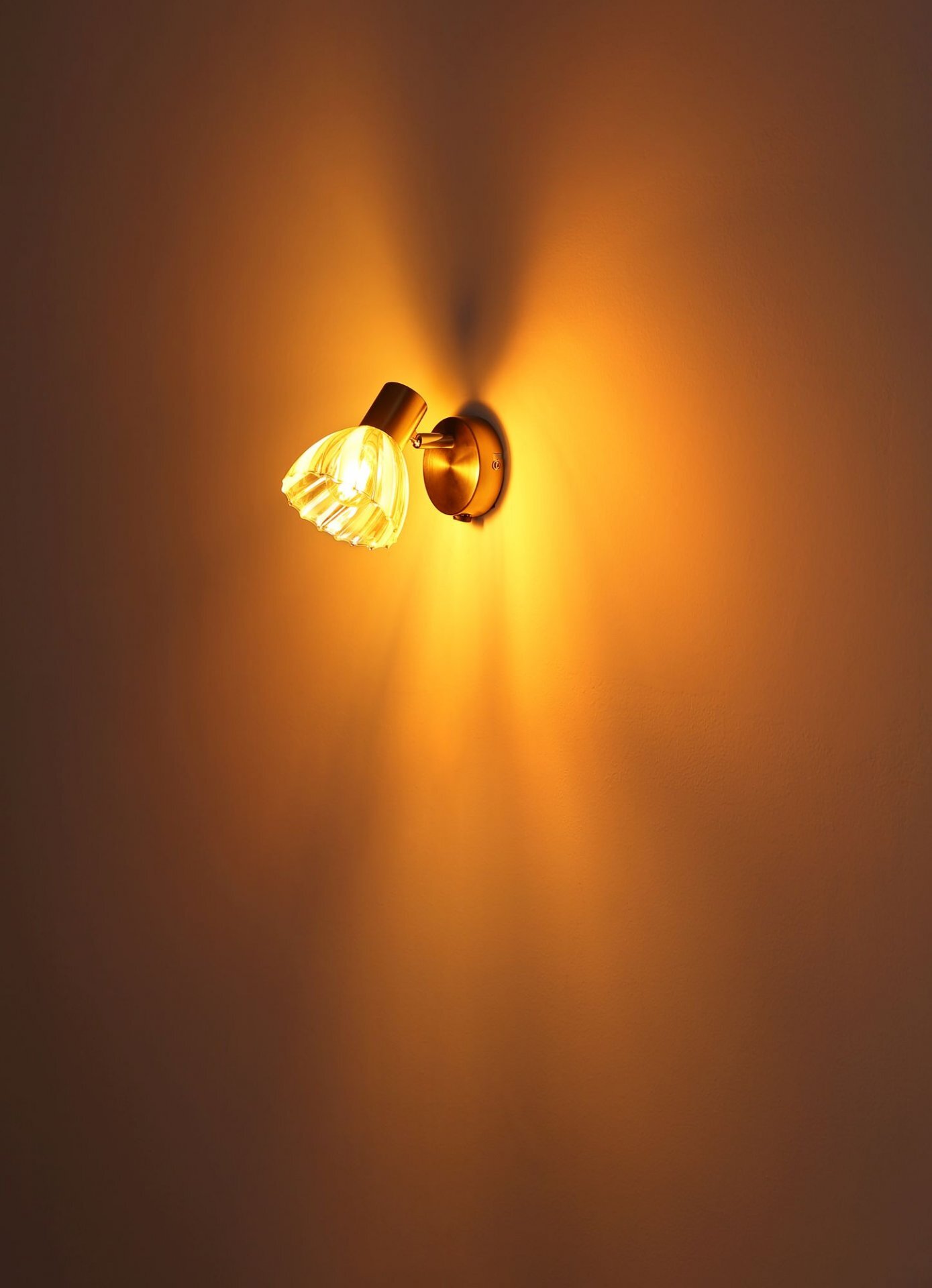 Na obrazie widoczna jest lampa reflektorowa emitująca ciepłe, żółte światło, zamontowana na ścianie. Światło tworzy rozproszone promienie na powierzchni ściany.