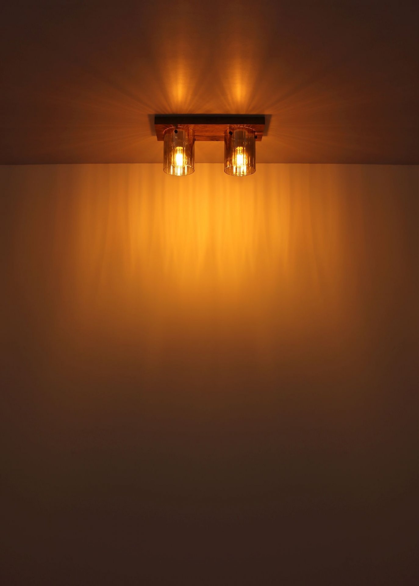 Na obrazie widoczna jest lampa sufitowa emitująca ciepłe, żółte światło, które rozprasza się na suficie i ścianie. Lampa ma dwa źródła światła zamontowane na prostokątnej podstawie.