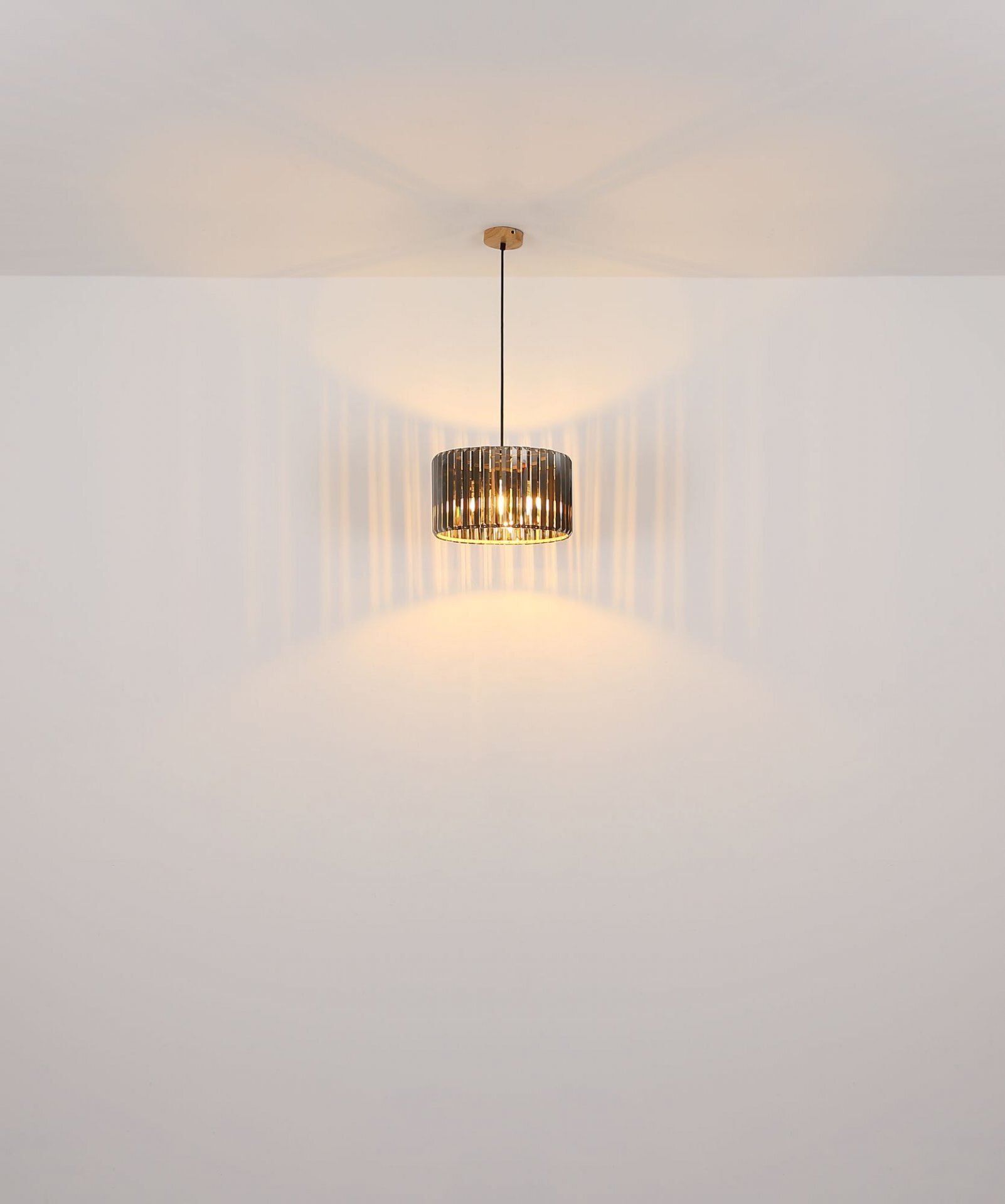 Na obrazie widoczna jest wisząca lampa z ciemnym, ażurowym kloszem, emitująca ciepłe światło na ścianę i sufit. Lampa jest zawieszona na cienkim przewodzie, a jej światło tworzy wzory na otaczających powierzchniach.