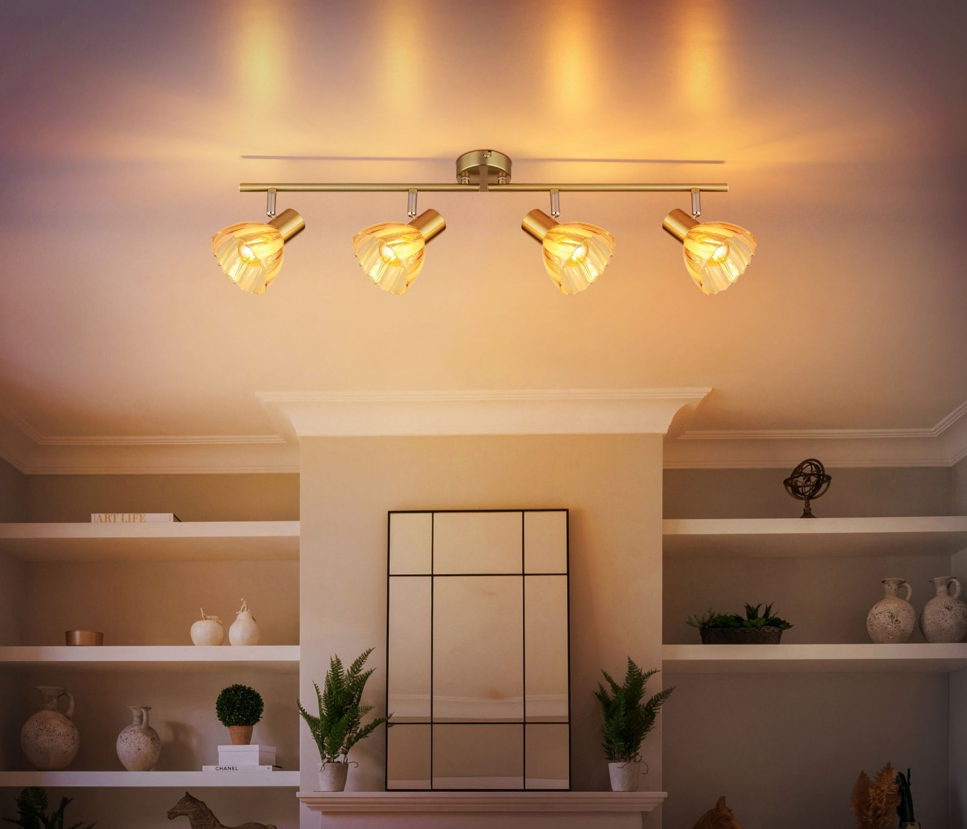Na suficie zamontowana jest lampa z czterema regulowanymi reflektorami, emitująca ciepłe światło. W tle widoczne są półki z dekoracjami i lustro na ścianie.