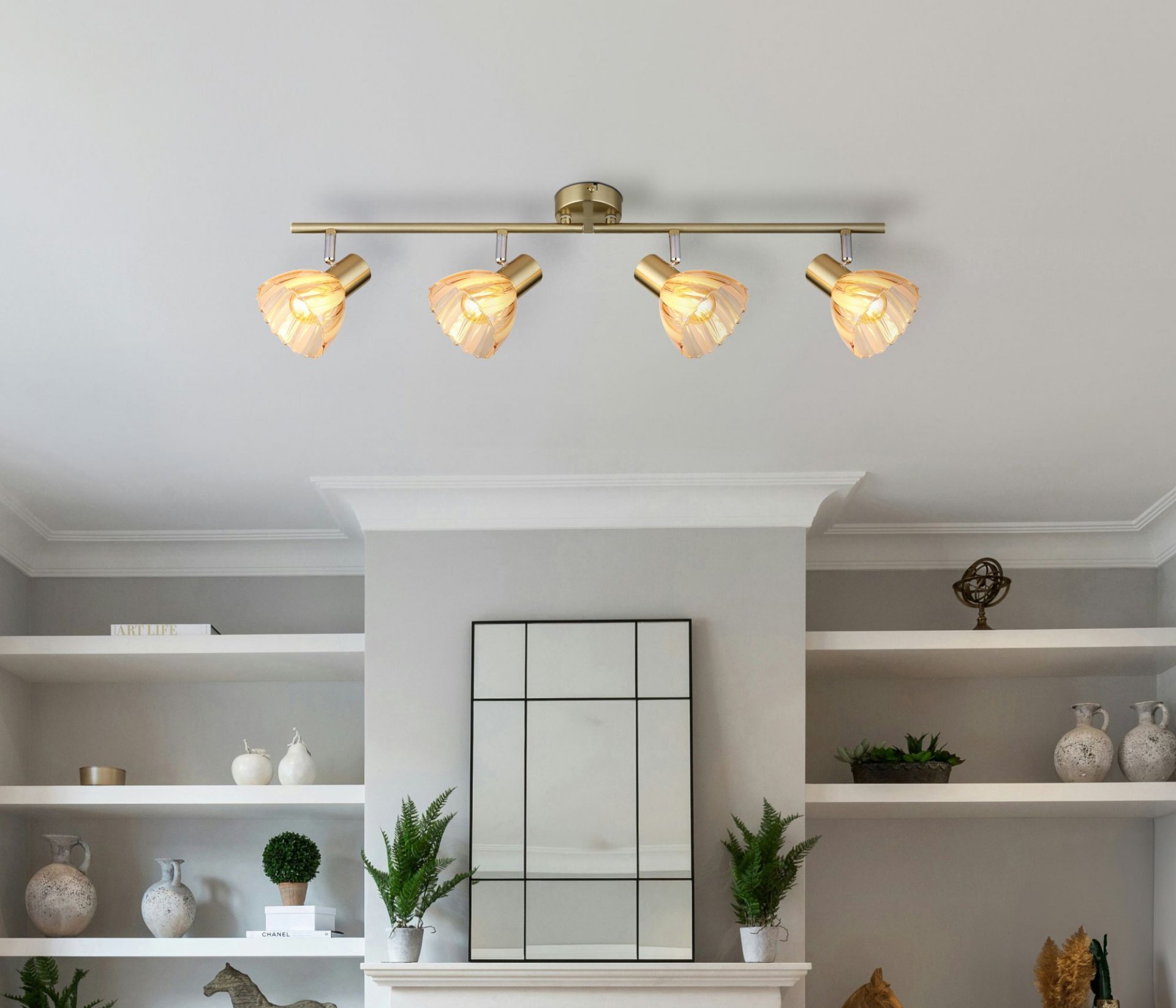 Na suficie zamontowana jest lampa z czterema regulowanymi reflektorami o nowoczesnym designie. W tle widoczne są półki z dekoracjami i roślinami oraz lustro na ścianie.
