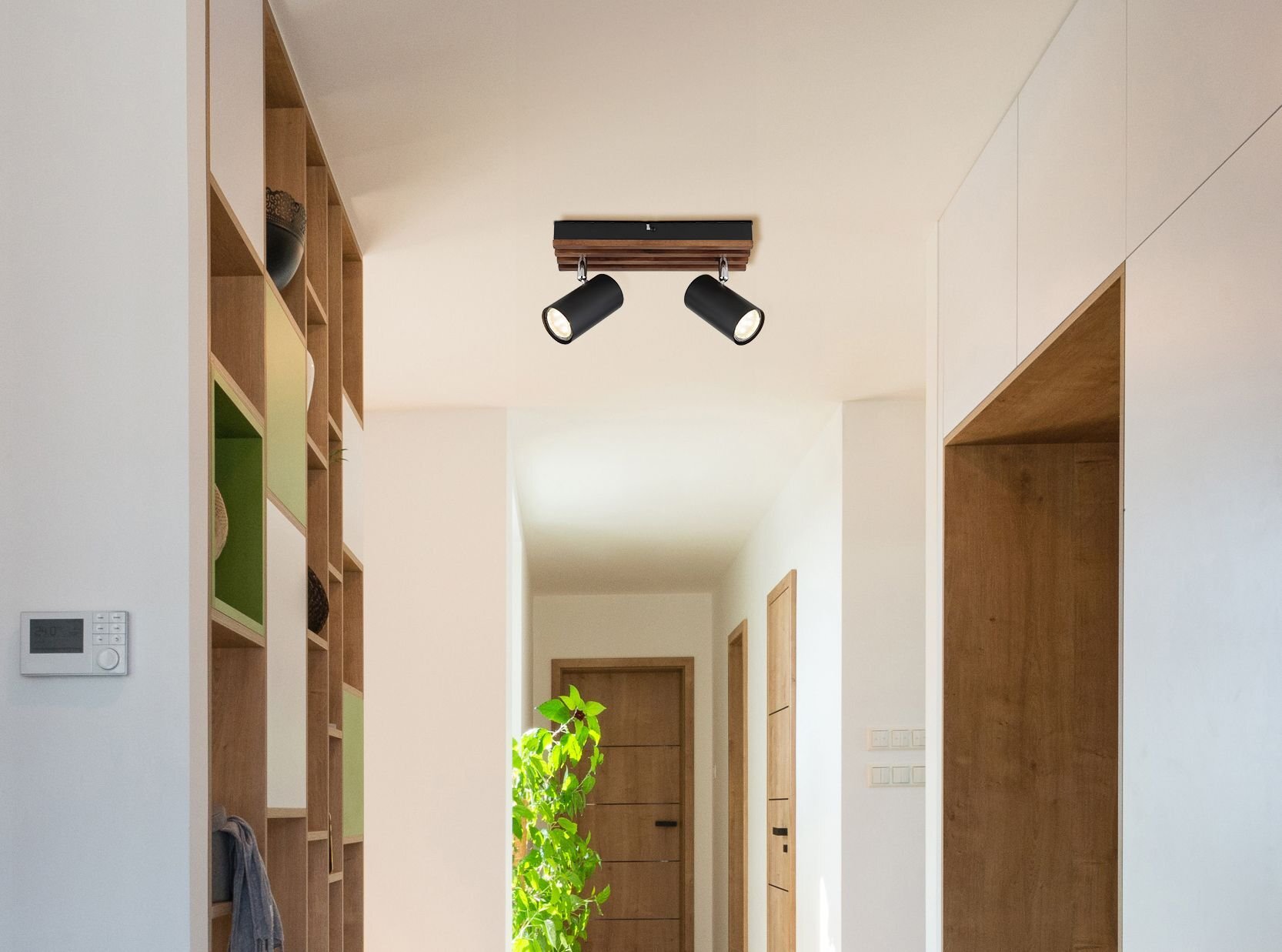 Na suficie znajduje się nowoczesna lampa sufitowa z dwoma regulowanymi reflektorami w czarnym kolorze. Wnętrze jest jasne, z drewnianymi drzwiami i półkami oraz rośliną w doniczce.