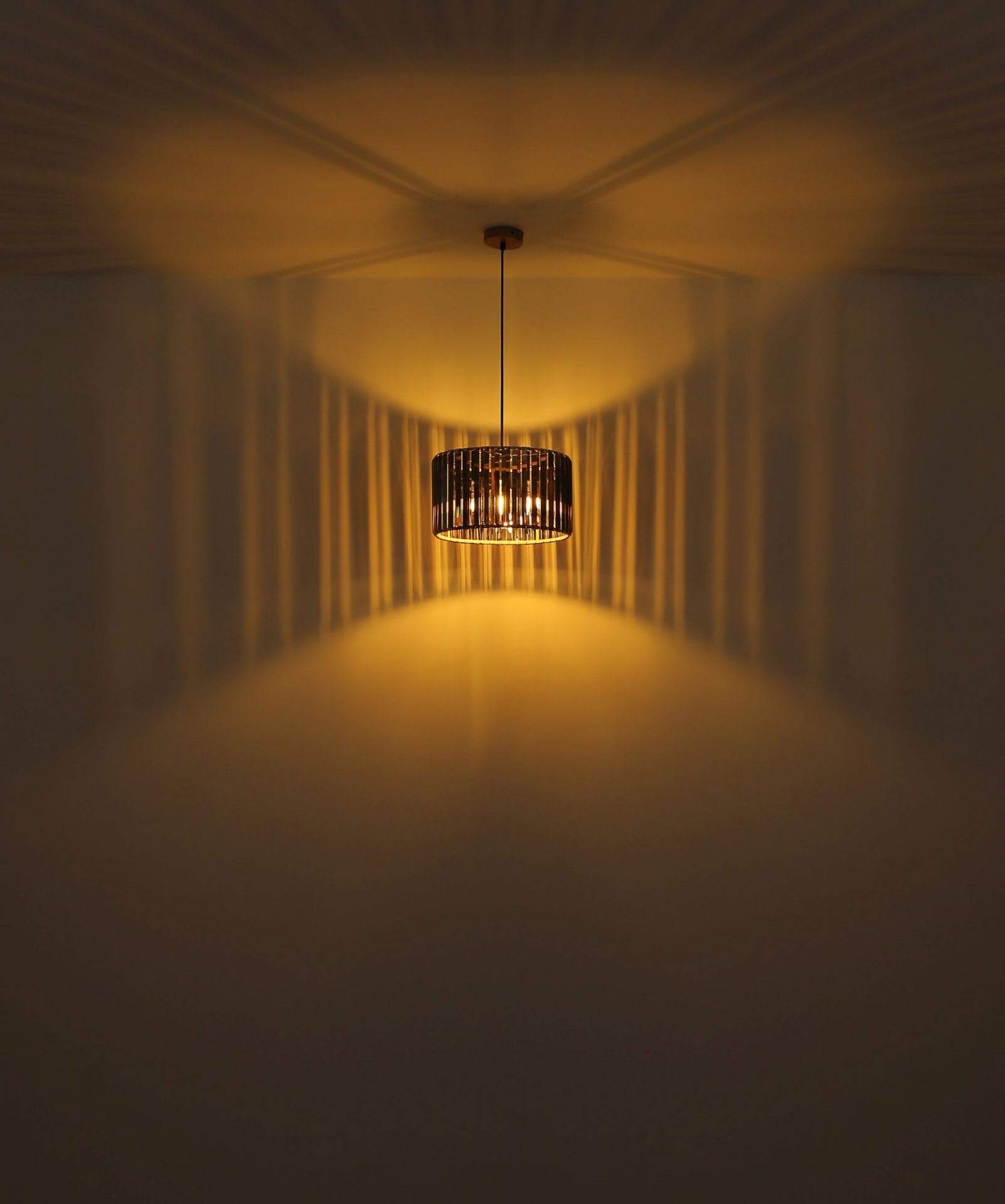 Na obrazie widoczna jest wisząca lampa z ciemnym kloszem, która emituje ciepłe, żółte światło, tworząc wzory na ścianie i suficie. Światło rozchodzi się w formie promieni, tworząc efektowne refleksy.