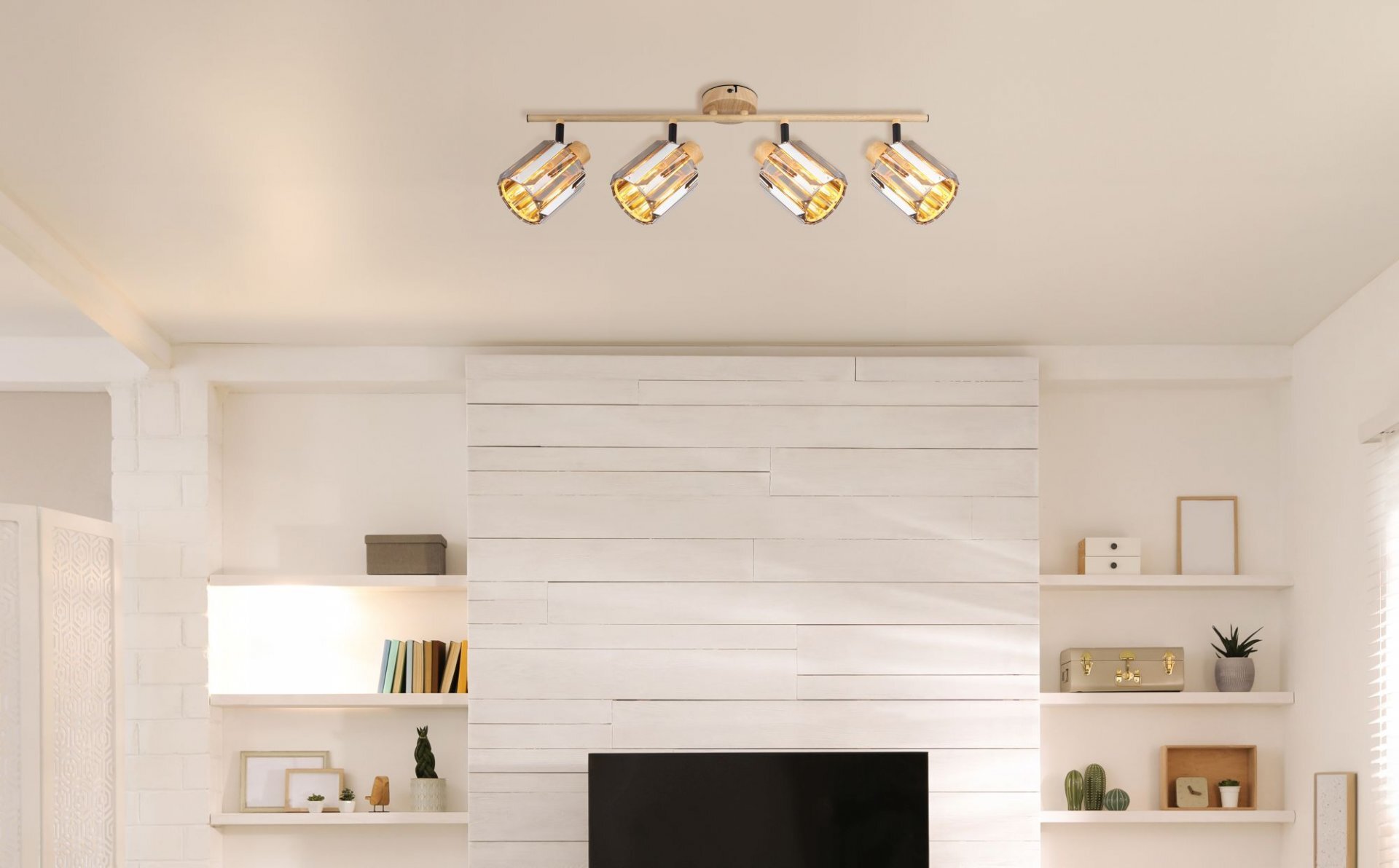Na suficie zamontowana jest lampa reflektorowa z czterema punktami świetlnymi. W tle widać jasną ścianę z półkami, na których znajdują się książki i dekoracje.