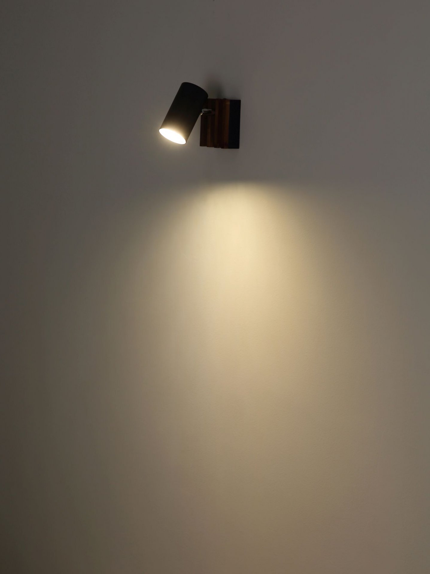 Na ścianie zamontowana jest czarna lampa reflektorowa, emitująca skupione światło na powierzchnię poniżej.