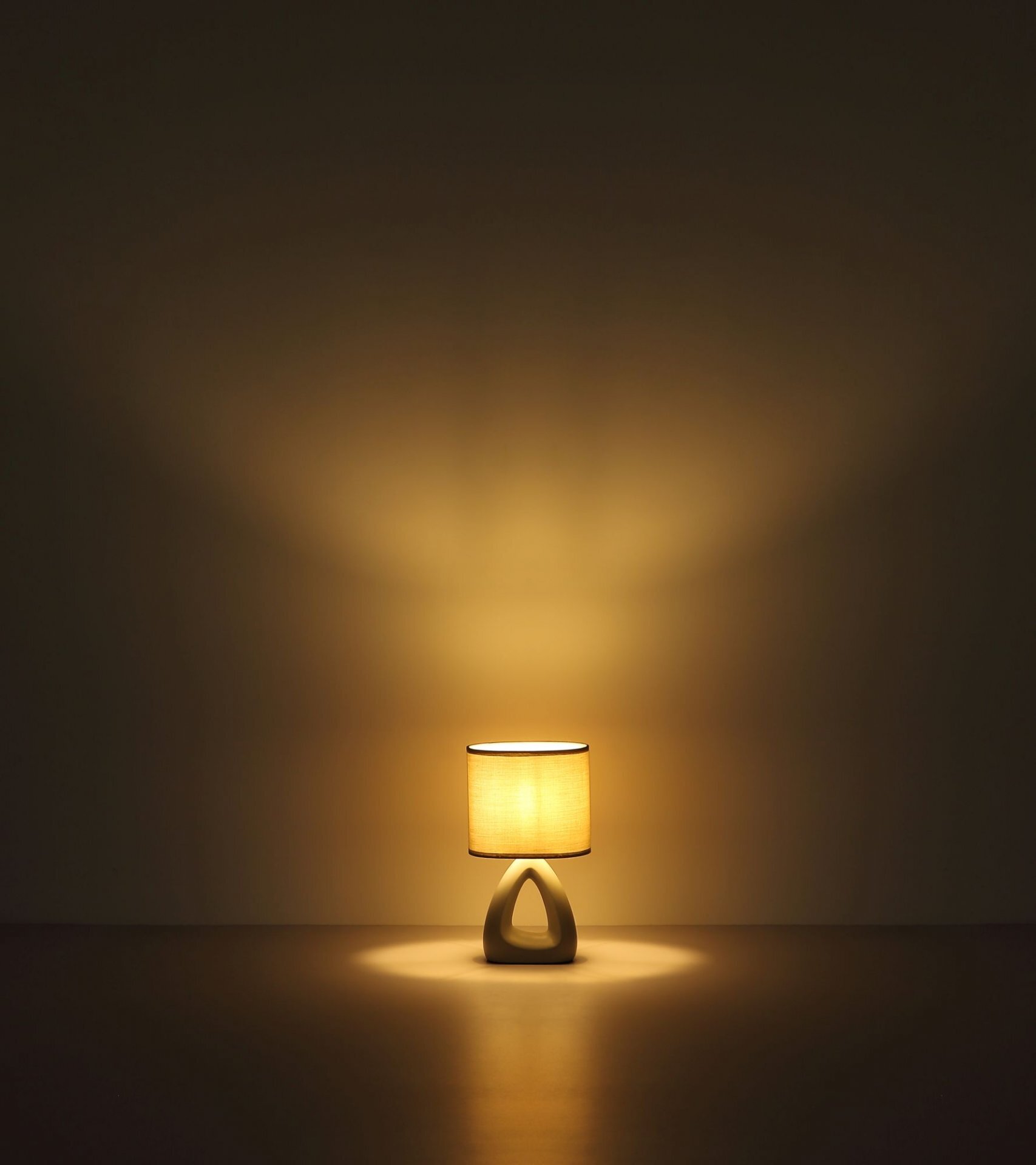 Na obrazie widoczna jest nowoczesna lampa stołowa z abażurem, emitująca ciepłe, rozproszone światło na ścianę i podłogę. Podstawa lampy ma geometryczny kształt przypominający trójkąt.