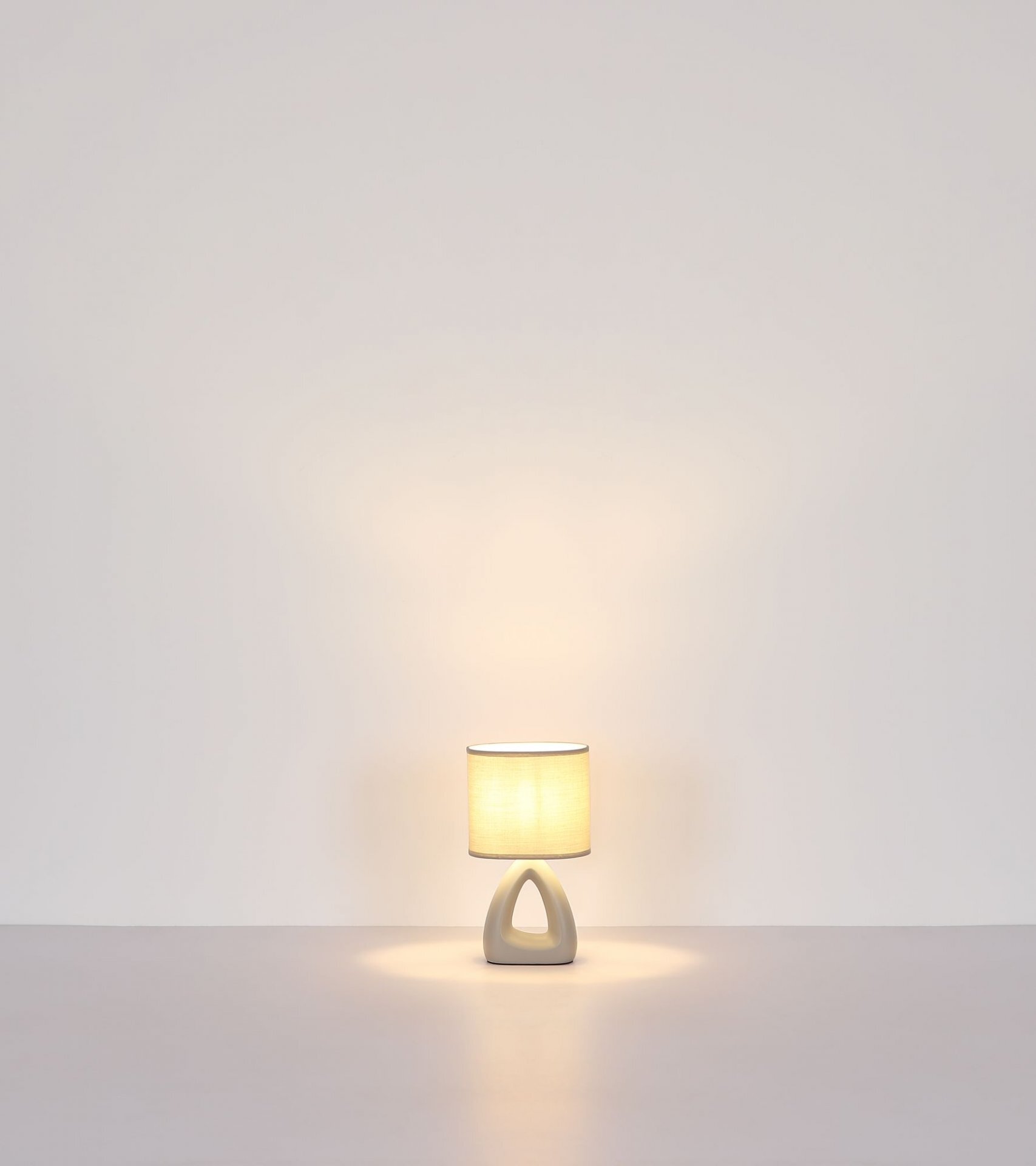 Na obrazie widoczna jest nowoczesna lampa stołowa z abażurem w kształcie walca, emitująca ciepłe światło. Podstawa lampy ma geometryczny kształt z otworem pośrodku.