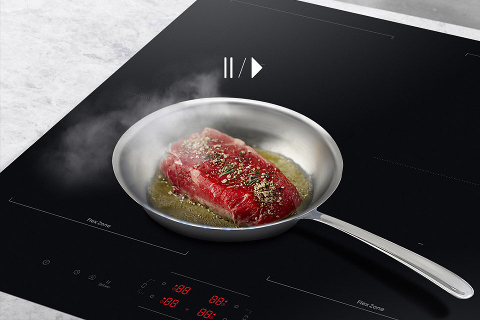 Na płycie indukcyjnej znajduje się patelnia z kawałkiem mięsa, z której unosi się para. Na płycie widoczny jest napis \'Flex Zone\' oraz cyfry \'88 88\'.