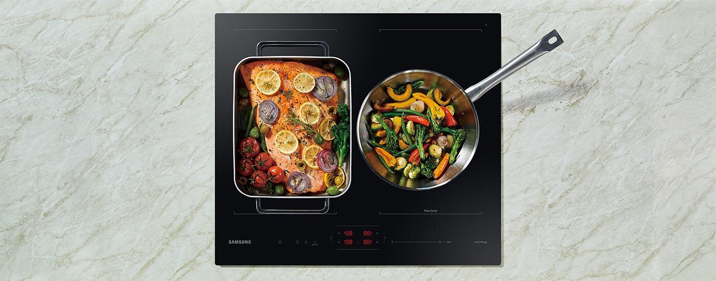 Na płycie indukcyjnej znajdują się dwa naczynia z jedzeniem: jedno z pieczonym łososiem i cytryną, drugie z mieszanką warzyw. Na płycie widoczne jest logo \'SAMSUNG\' oraz panel dotykowy z czerwonymi wskaźnikami.