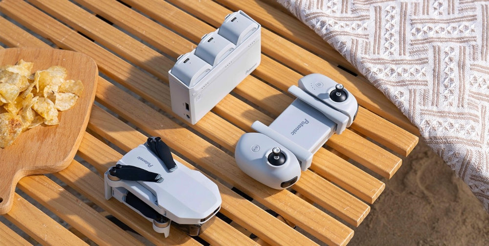 Na drewnianym stole leży złożony dron, kontroler oraz ładowarka z trzema miejscami na baterie, wszystkie w jasnym kolorze. Obok znajduje się deska z chipsami i fragment wzorzystego materiału.