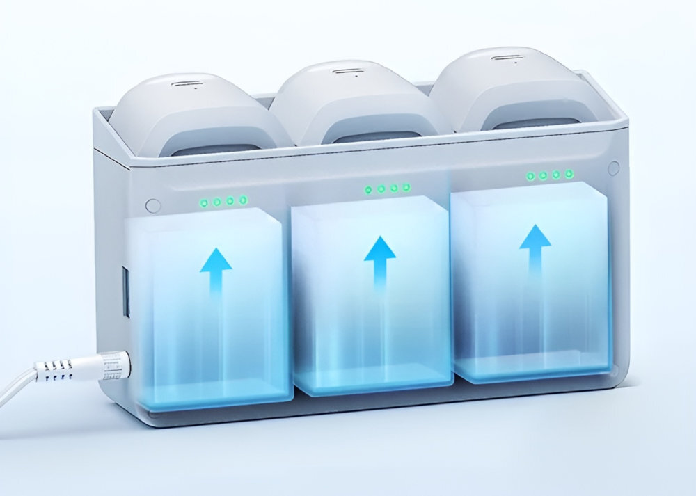 Na obrazie widoczna jest stacja ładująca z trzema miejscami na baterie, z niebieskimi strzałkami wskazującymi do góry. Na górze każdego miejsca znajdują się zielone diody sygnalizacyjne.