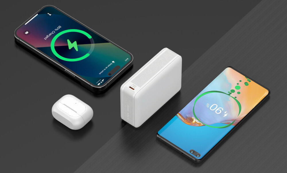 Na obrazie widoczny jest biały powerbank stojący pionowo obok dwóch smartfonów z widocznymi ekranami ładowania oraz białych słuchawek bezprzewodowych w etui. Na ekranach smartfonów widoczne są ikony ładowania i procenty naładowania baterii.