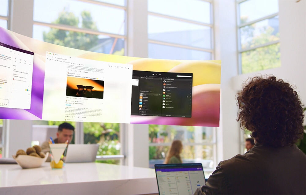 Osoba siedzi przy biurku z laptopem, patrząc na wirtualny ekran z kilkoma otwartymi oknami aplikacji. W tle widać dużą przeszkloną ścianę i inne osoby pracujące przy biurkach.