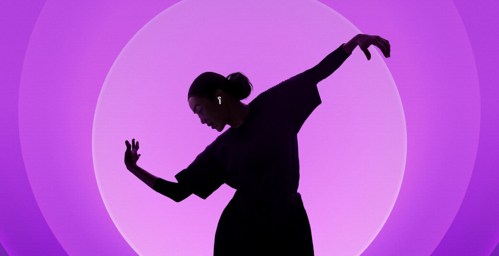 Osoba w ciemnym ubraniu tańczy na tle fioletowego, okrągłego światła. W uchu widoczne są białe słuchawki douszne.