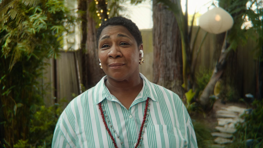 Osoba stoi na zewnątrz w ogrodzie, nosząc słuchawki douszne. W tle widoczne są drzewa i rośliny.