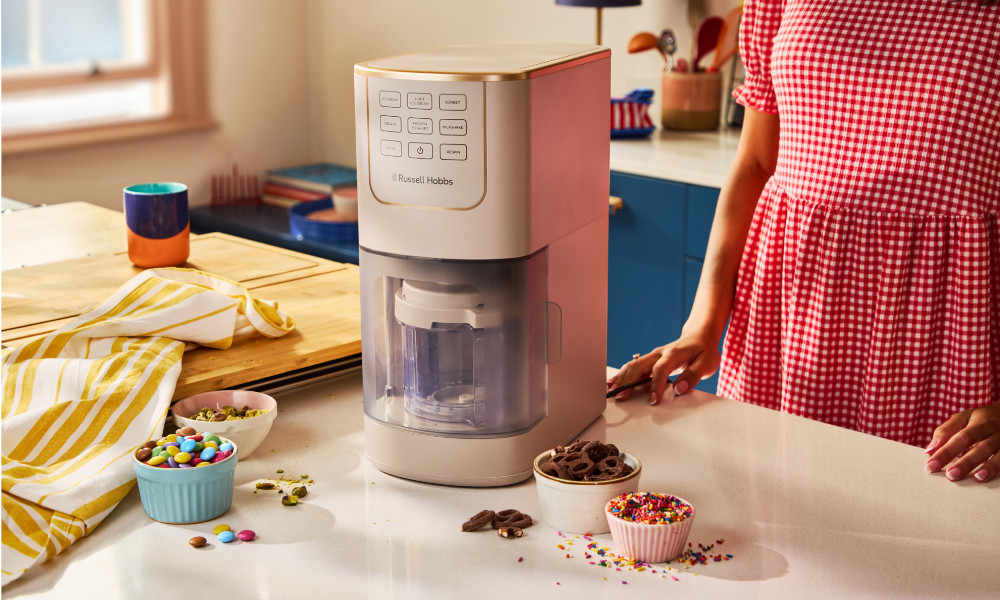 Na blacie kuchennym stoi maszynka do lodów z widocznym logo \'Russell Hobbs\'. Obok znajdują się miseczki z kolorowymi posypkami i czekoladą.
