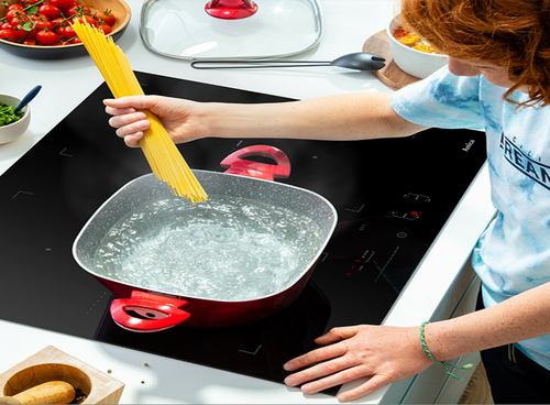 Osoba gotuje makaron w czerwonym garnku na czarnej płycie indukcyjnej z widocznym logo \'Amica\'. W tle znajdują się różne kuchenne akcesoria i składniki.