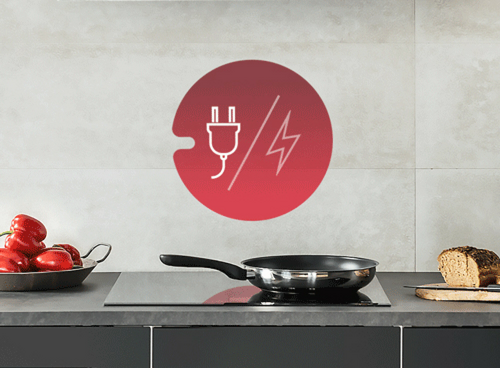 Na blacie kuchennym znajduje się płyta indukcyjna z patelnią. Obok leży chleb i miska z czerwonymi paprykami, a na ścianie widoczna jest ikona z wtyczką i symbolem błyskawicy na czerwonym tle.