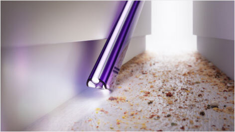 Fioletowa końcówka odkurzacza wąska, czyszcząca przestrzeń między dwoma powierzchniami, z widocznymi drobinkami kurzu i brudu.