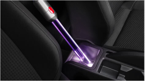 Na zdjęciu widać końcówkę odkurzacza z fioletowym światłem, używaną do czyszczenia wnętrza samochodu, w szczególności przestrzeni między siedzeniami. W tle widoczne są elementy wnętrza samochodu, takie jak uchwyty na kubki.