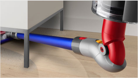 Na obrazie widoczna jest końcówka odkurzacza w kolorach czerwonym, szarym i niebieskim, czyszcząca podłogę pod meblem. W tle widać fragment podłogi i ściany.