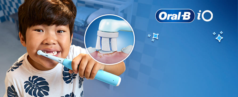 Dziecko trzyma niebieską szczoteczkę elektryczną Oral-B, a obok widoczna jest ilustracja końcówki szczoteczki na tle dziąseł. W prawym górnym rogu znajduje się logo \'Oral-B iO\'.