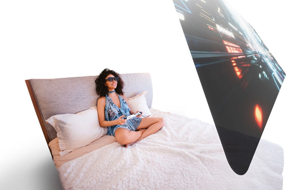Okulary XREAL One Pro prezentacja okularów podczas użytkowania kobieta siedząca na łóżku grająca na padzie z założonymi okularami przed nią wirtualny ekran białe tło cztery tryby pracy rozszerzenie pola widzenia śledzenie otoczenia i treści AR komfort w ruchu przypięcie ekranu podążanie za ruchami głowy stabilność