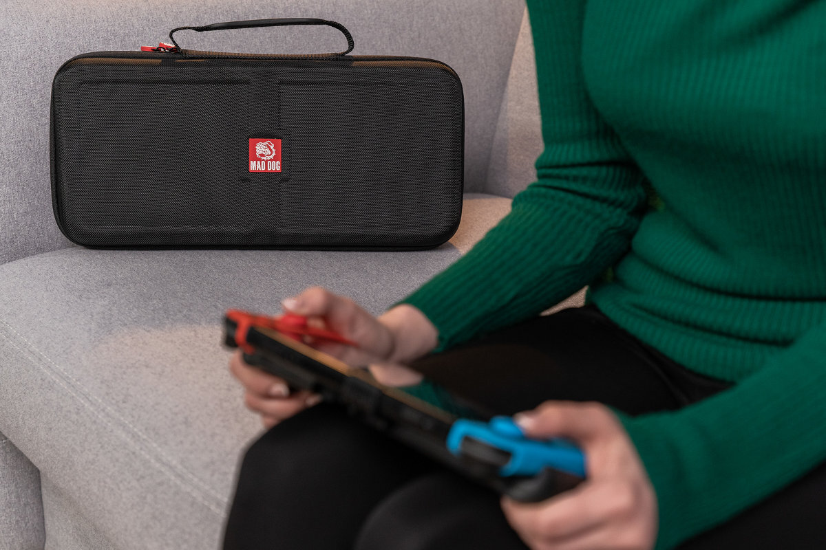 Na szarej kanapie leży czarne etui z logo \'MAD DOG\'. Obok osoba w zielonym swetrze trzyma konsolę do gier.