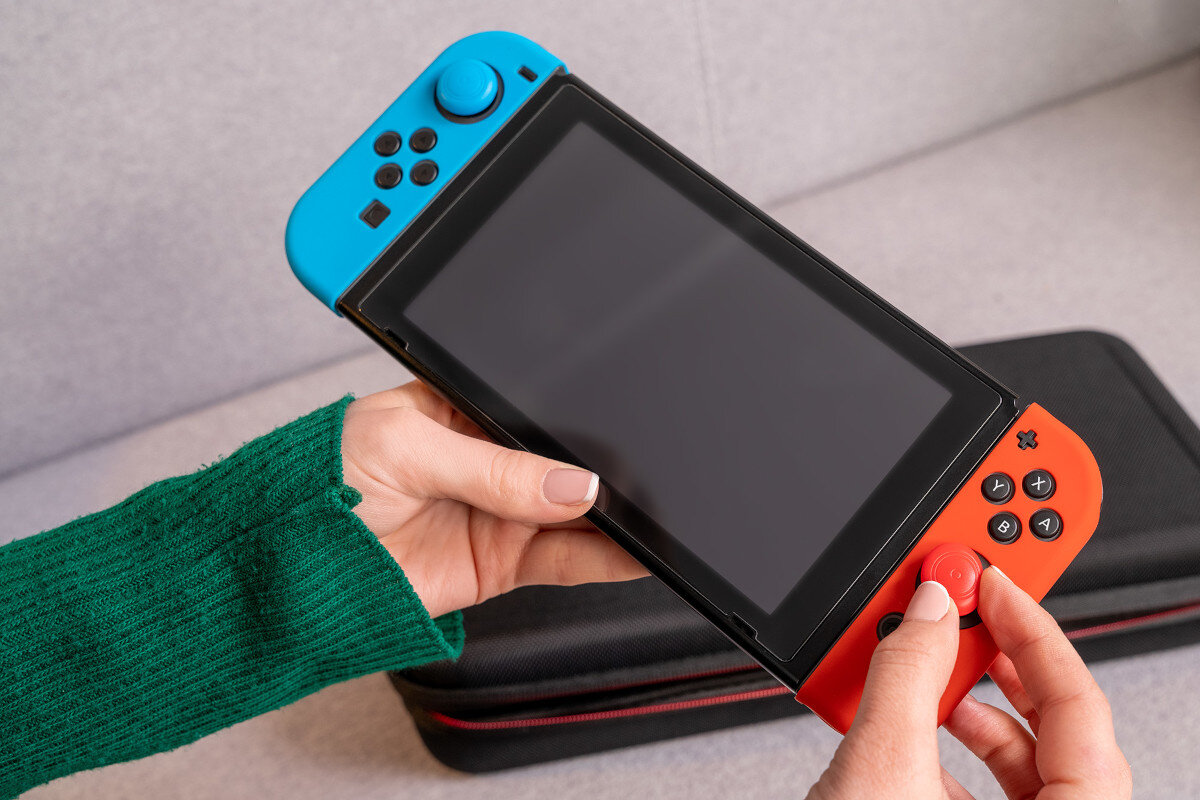 Osoba trzyma przenośną konsolę do gier z niebieskim i czerwonym kontrolerem. W tle widoczne jest czarne etui z czerwonym zamkiem błyskawicznym.