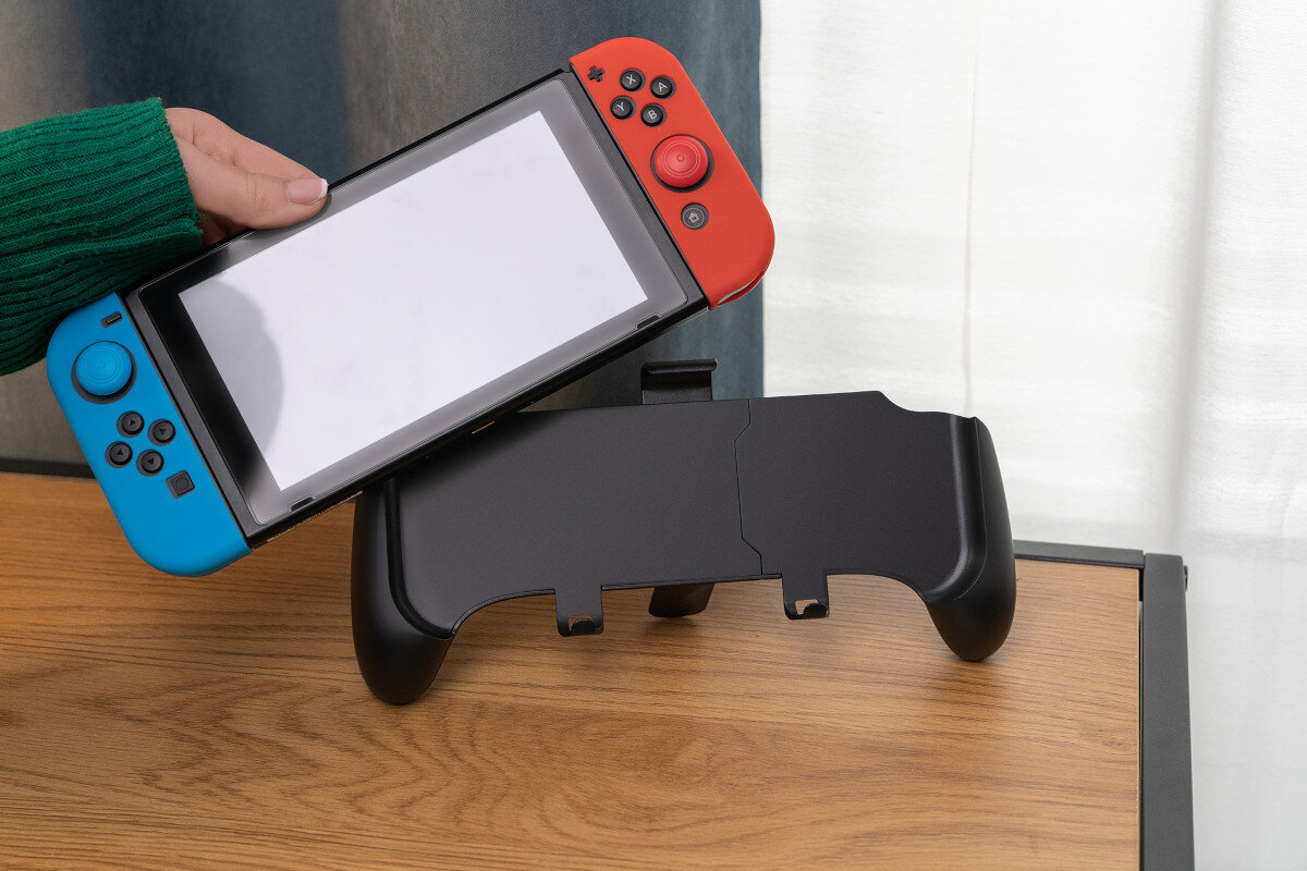 Osoba trzyma konsolę do gier z czerwonym i niebieskim kontrolerem, obok której znajduje się czarne etui na drewnianym stole.