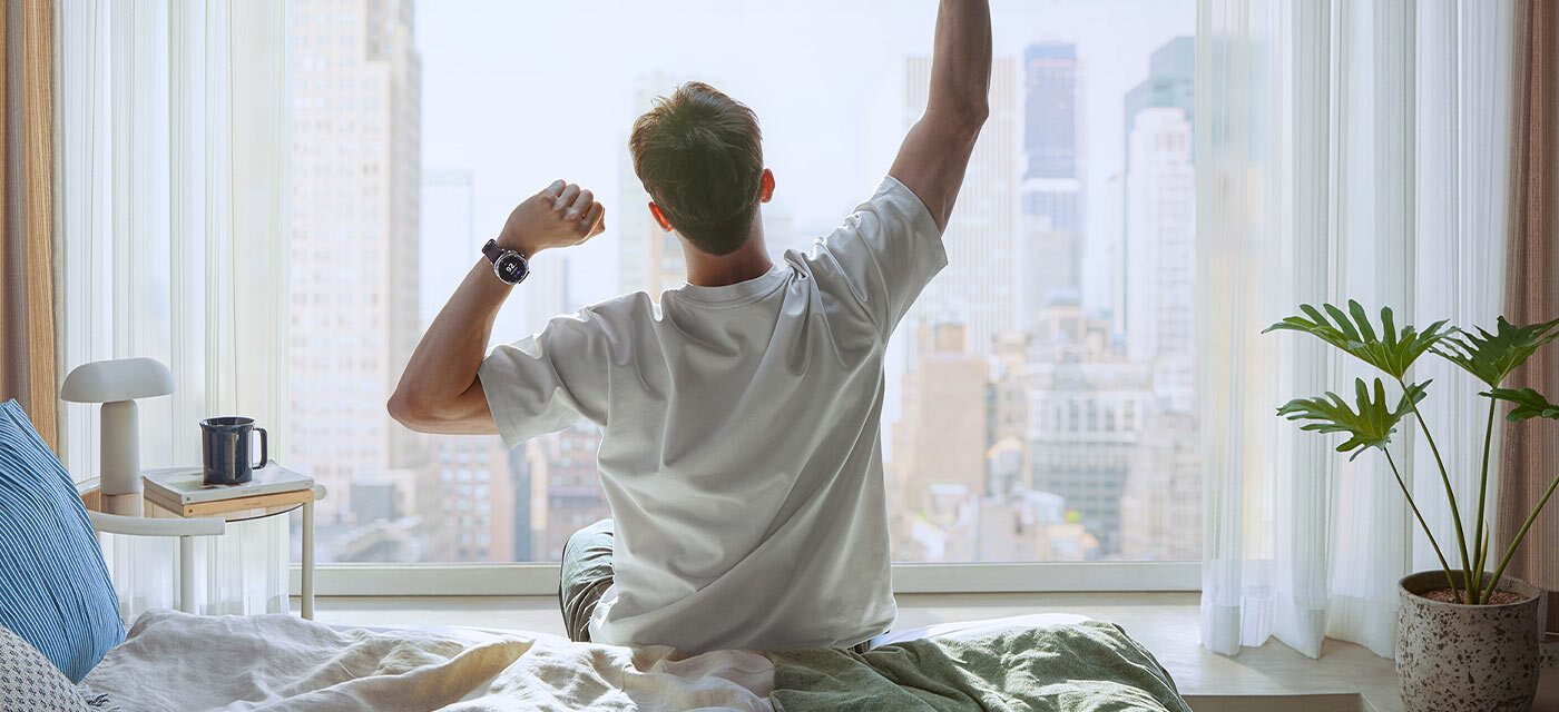 Osoba siedzi na łóżku w jasnym pokoju, patrząc przez duże okno na panoramę miasta. Na nadgarstku ma założony smartwatch.