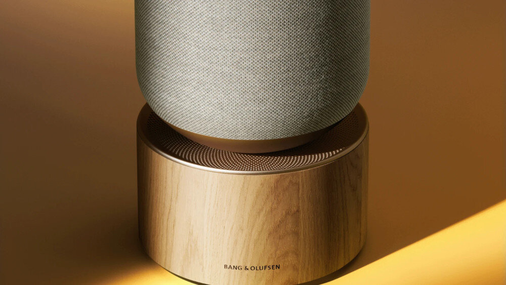Głośnik o cylindrycznym kształcie z tekstylną górną częścią i drewnianą podstawą. Na podstawie widoczne jest logo \'BANG & OLUFSEN\'.