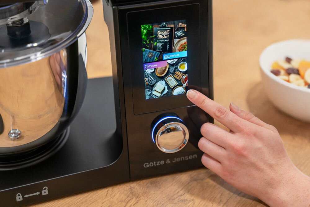 Robot kuchenny z kolorowym ekranem dotykowym, na którym widoczne są opcje \'Modes\' i \'Recipes\'. Obok znajduje się miska z pokrojonymi owocami, a na urządzeniu widnieje napis \'Götze & Jensen\'.