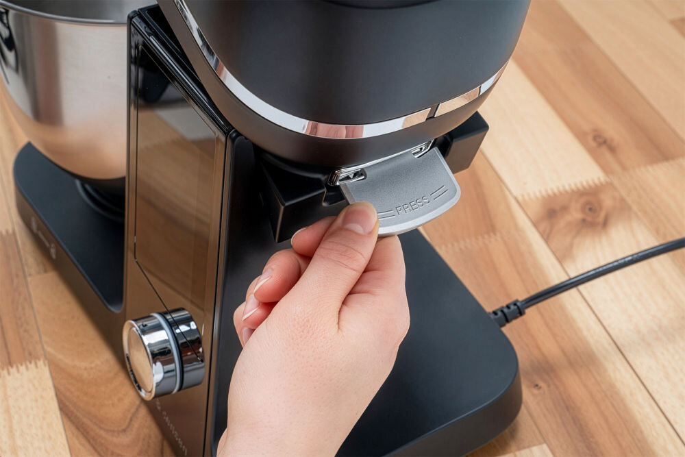 Osoba trzyma element robota kuchennego planetarnego, na którym widnieje napis \'PRESS\'. Urządzenie stoi na drewnianym blacie i jest podłączone do prądu.