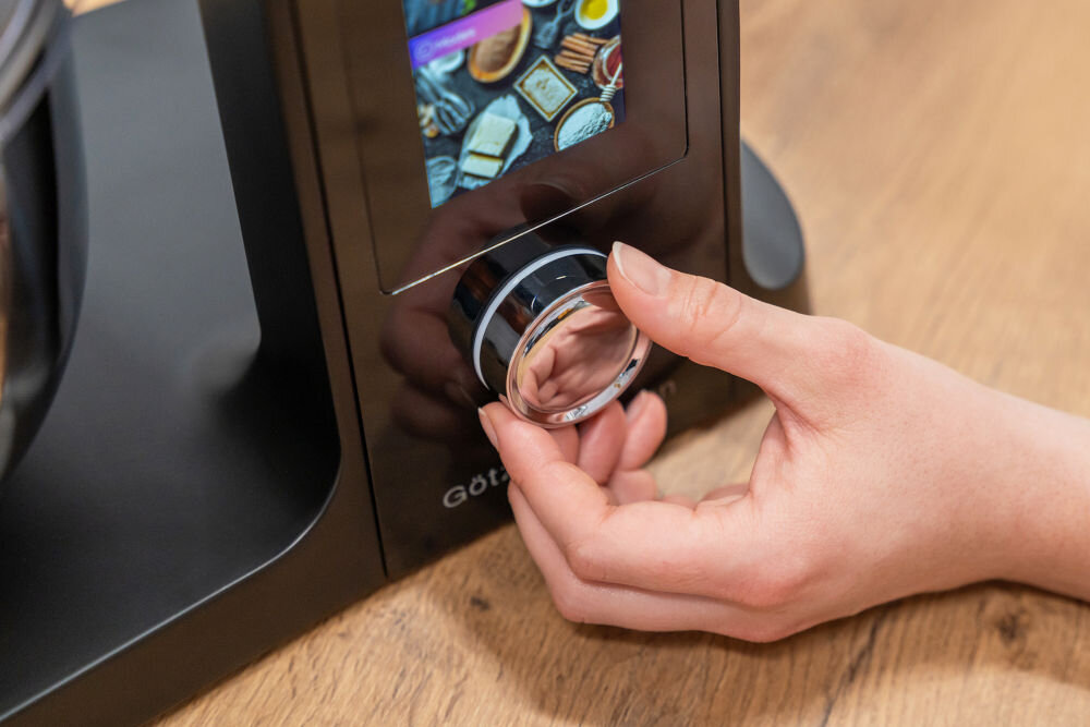 Osoba obsługuje pokrętło na panelu sterowania robota kuchennego z kolorowym ekranem wyświetlającym różne składniki. Na urządzeniu widoczne jest logo \'Götze & Jensen\'.